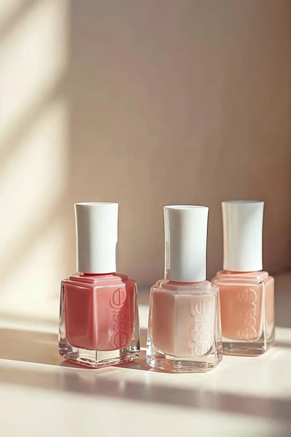 Three nail polish bottles in shades of coral pink and nude arranged on a light background, showcasing products for almond nai