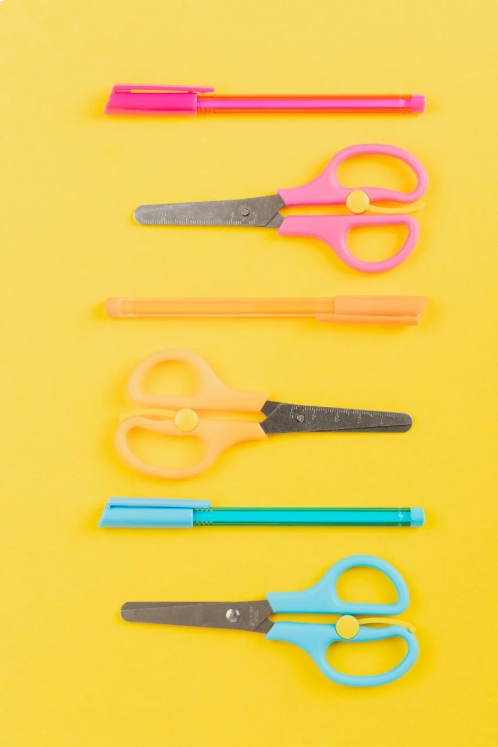 Flat lay of colorful school supplies including pink and orange scissors, pens, and a blue marker on a yellow background