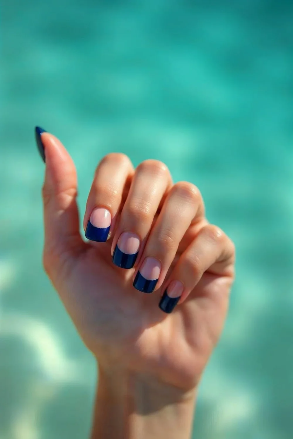 Navy blue French tips on medium-length round nails against turquoise water background showcasing beach nails