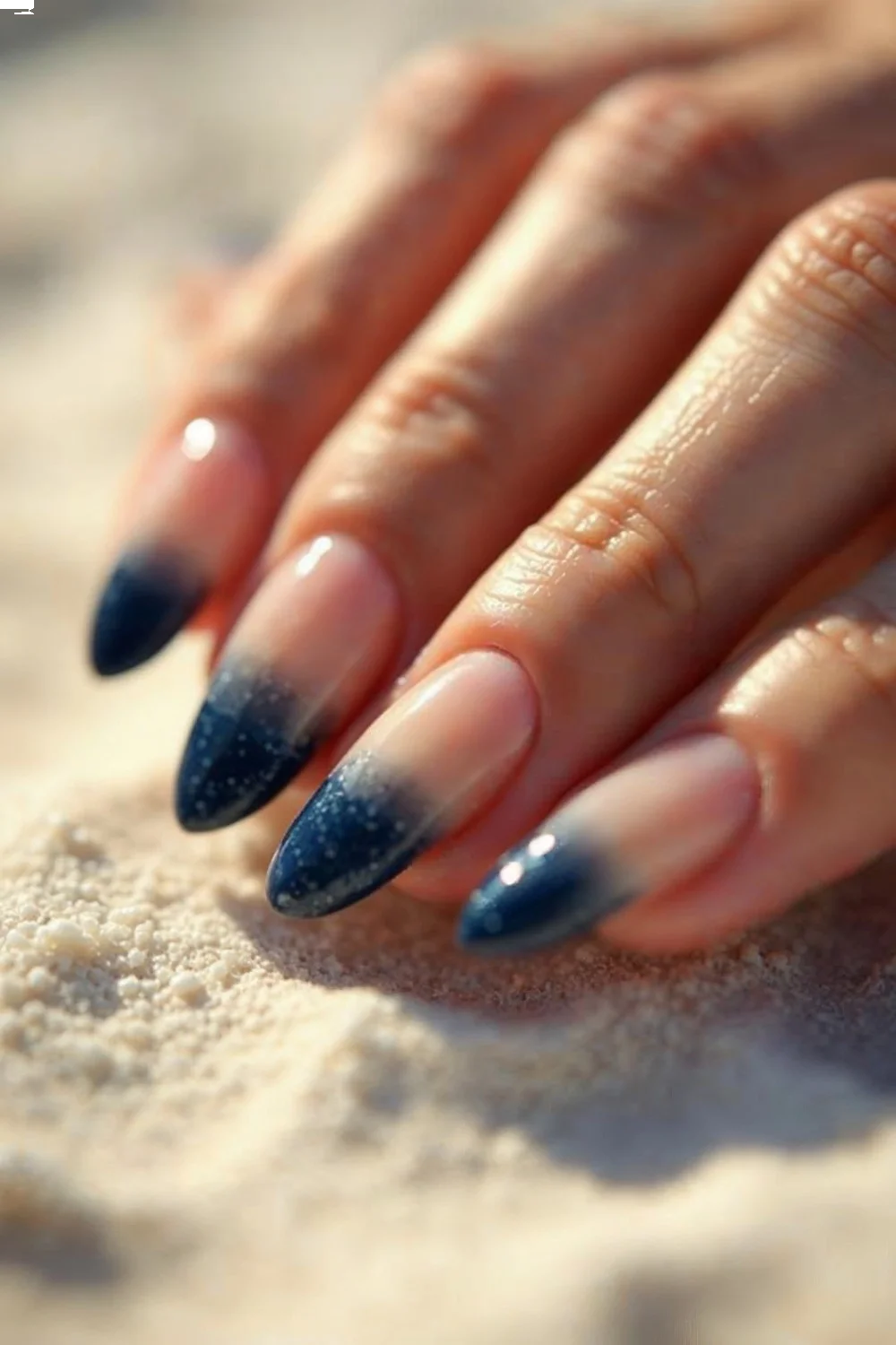 Almond-shaped beach nails with sandy beige to deep navy gradient on a white sand background