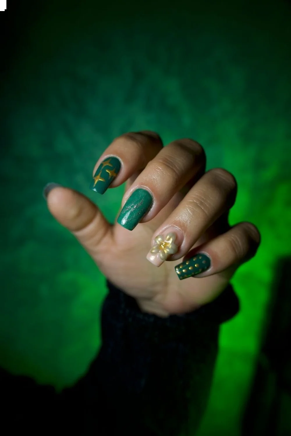 dark green nails with gold accents and a floral design on a hand against a green background