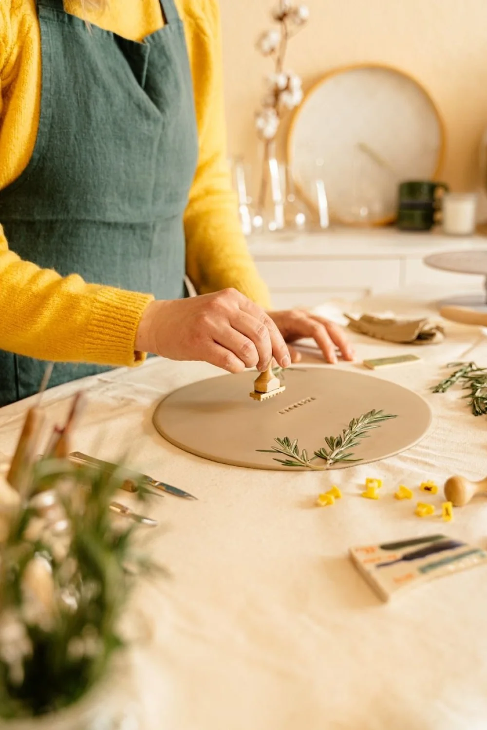 A person in a yellow sweater uses a stamper to transfer a design onto a nail, surrounded by nail art tools and greenery, illu