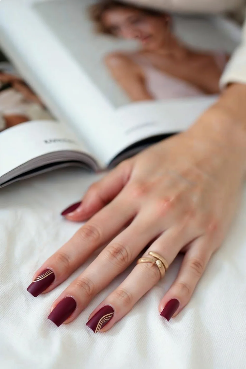 Burgundy matte nails with gold diagonal glossy stripes on a hand resting on a white surface next to a magazine