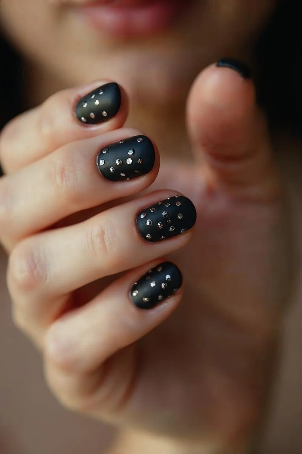 Matte black nails with glossy dots in a random pattern on a hand held up against a blurred background
