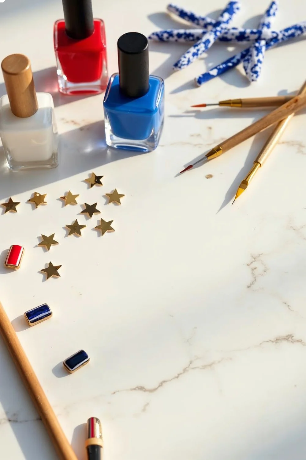 Red, white, and blue nail polishes with gold star decals, dotting tools, and striping brushes on a white marble surface for m