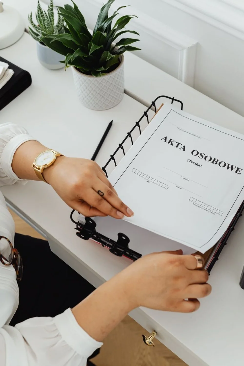 Hands holding a binder with a document titled "AKTA OSOBOWE" on a desk with a plant and stationery