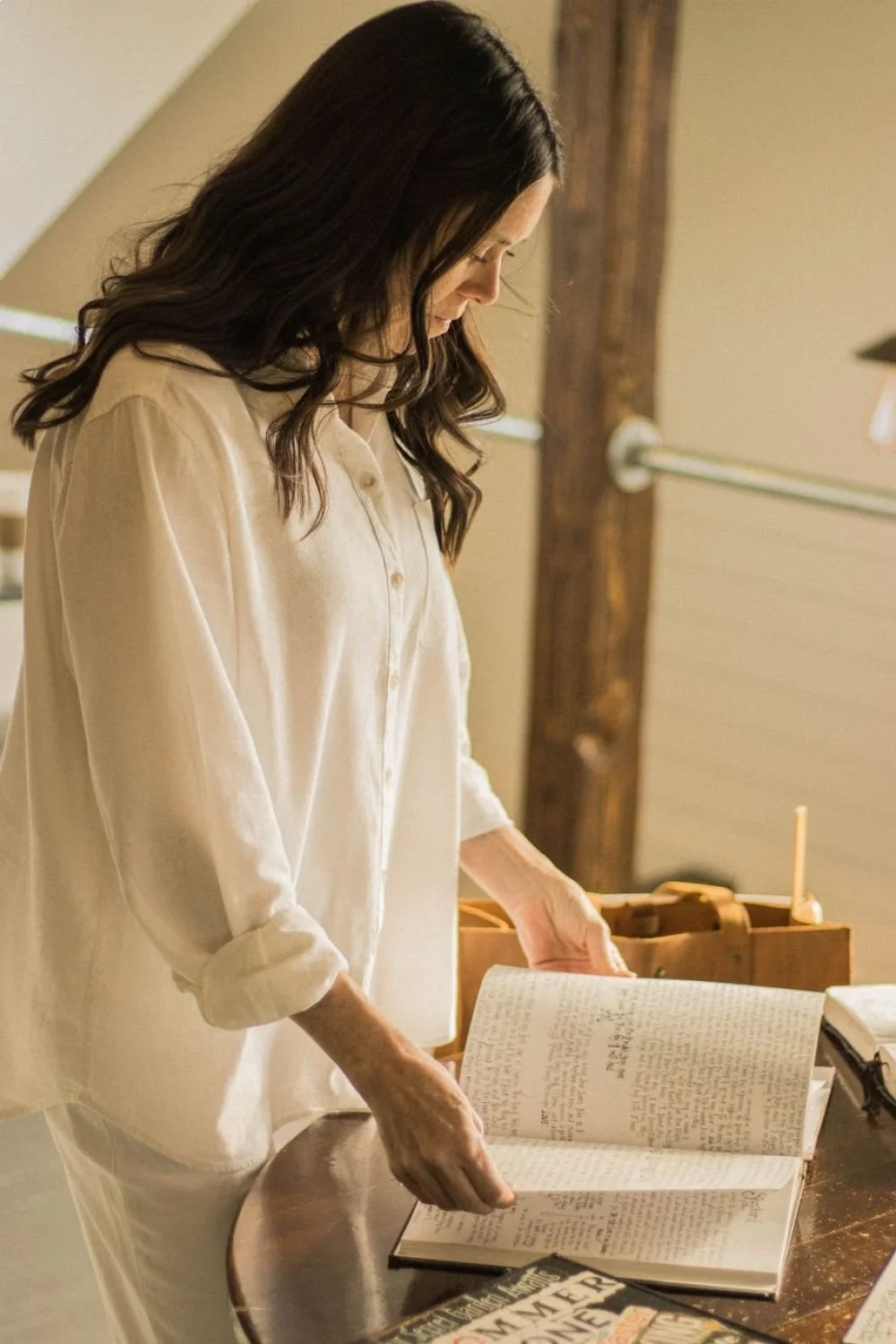 A woman in a white shirt stands at a table, reading a notebook with tropical nail designs visible on her nails