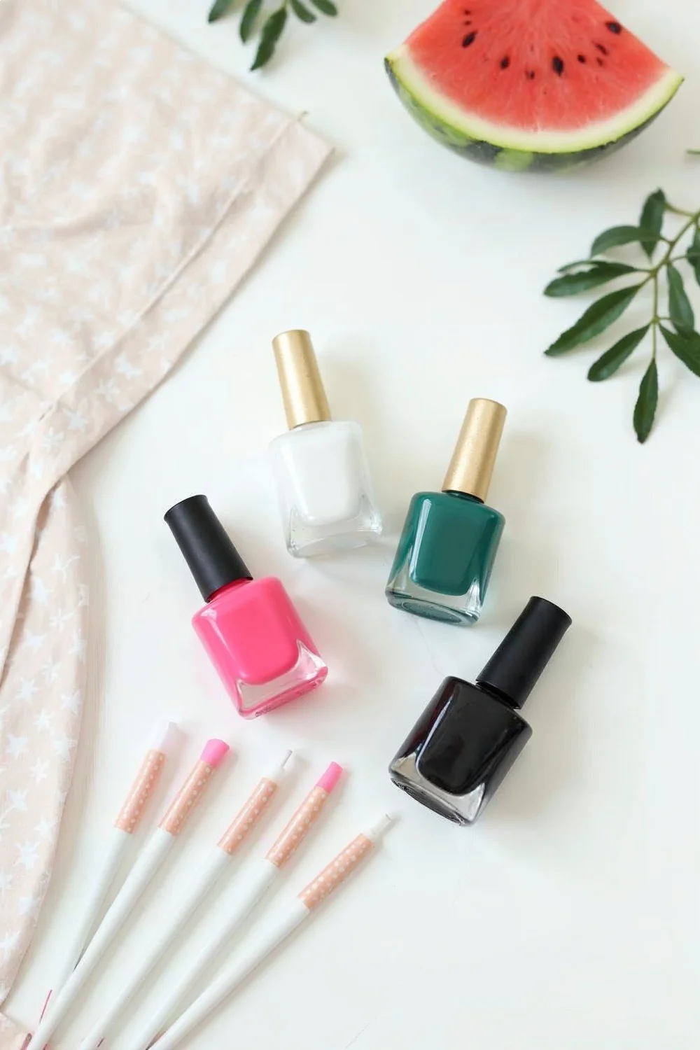 Flat lay of four nail polish bottles in pink, white, dark green, and black with dotting tools and a watermelon slice