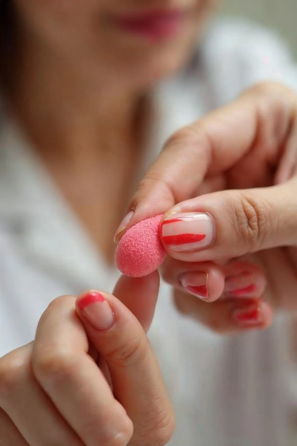 Hand holding a pink makeup sponge over coral pink nails with red stripes, demonstrating a bouncing motion for watermelon nail